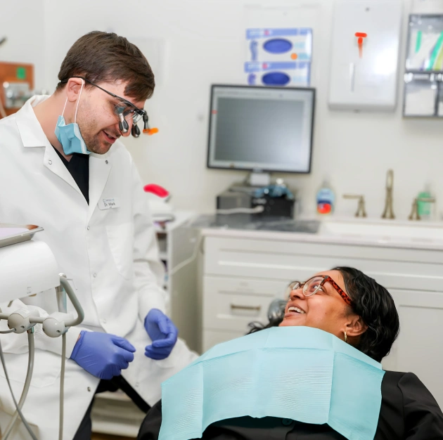 Dentist in light blue gloves examines smiling patient in dental treatment room.