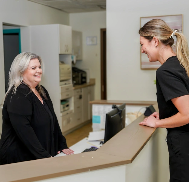 Two dental office staff members smiling at reception desk during patient consultation.